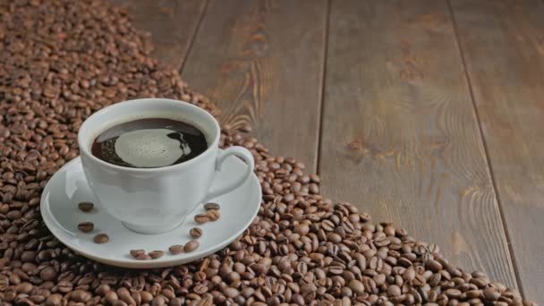 Kaukasische Hand, die schwarzen Kaffee in eine weiße Tasse mit sich drehenden Blasen auf einer flachen, teilweise mit gerösteten Kaffeebohnen bedeckten Holzoberfläche zurücklegt — Stockvideo