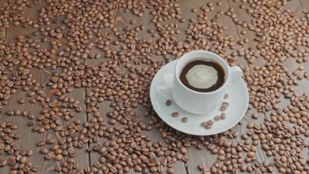 Café preto em uma caneca branca com bolhas de fiação em uma superfície plana de madeira parcialmente coberta com grãos de café torrados — Vídeo de Stock