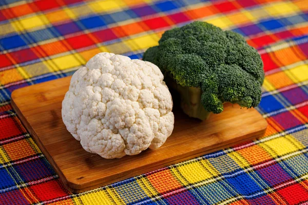 Una cabeza de brócoli y una cabeza de coliflor se encuentran en una tabla de cortar en la mesa con toalla de colores debajo —  Fotos de Stock