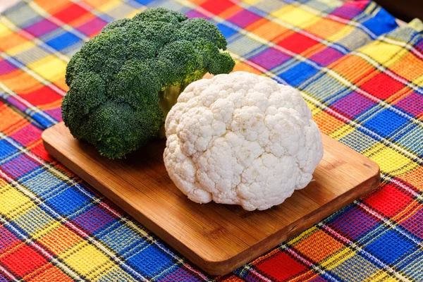 A head of broccoli and a head of cauliflower lie on a cutting board on the table with colorful towel underneath — Stockfoto