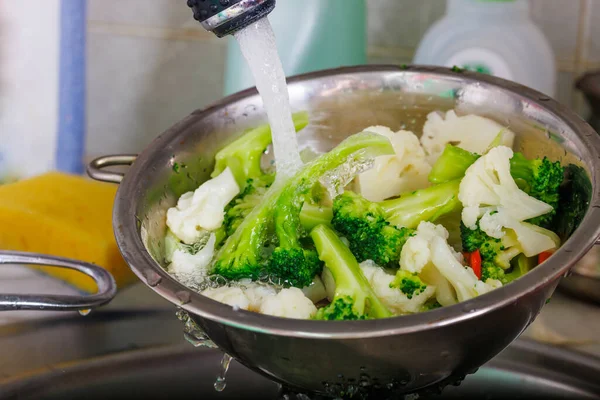 Agua del grifo fluye sobre la coliflor picada y el brócoli en un colador por encima del fregadero mientras enjuaga las verduras — Foto de Stock