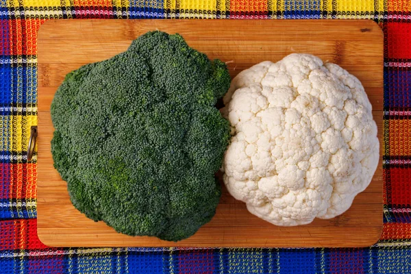 Una cabeza de brócoli y una cabeza de coliflor se encuentran en una tabla de cortar en la mesa con toalla de colores debajo — Foto de Stock
