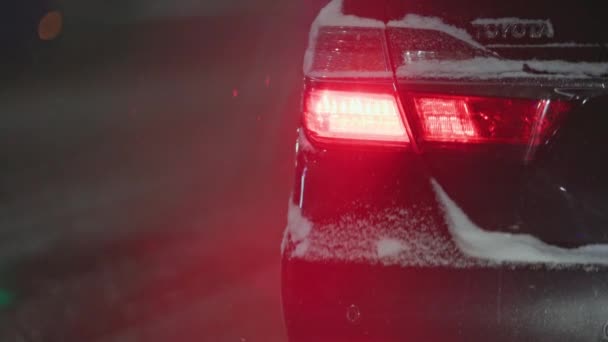 Κόκκινο φως ουρά του μαύρου αυτοκινήτου Toyota Camry στο δρόμο νύχτα στη χειμερινή χιονόπτωση — Αρχείο Βίντεο