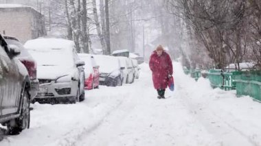 Kırmızı ceketli, yalnız, yaşlı bir Rus kadın park halindeki arabaların yanında, yoğun kar altında yürüyor.