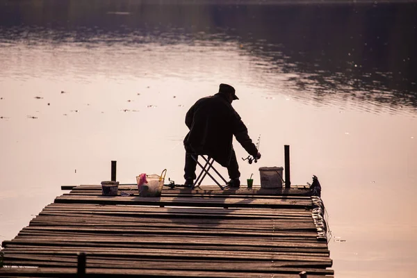 Silhouette of a fisherman from behind sitting on a wooden pier during evening fishing — 图库照片