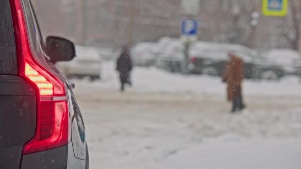 Fanale posteriore auto nera e pedoni sfocati attraversano la strada innevata in inverno nevicate di giorno — Video Stock