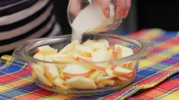 Пожилая женщина наливает сахар в стеклянную миску с нарезанными яблоками во время приготовления яблочного пирога — стоковое видео