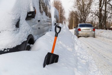 Plastic snow shovel in front of snow-covered minivan at sunny winter morning and silver car passing by in blurry background
