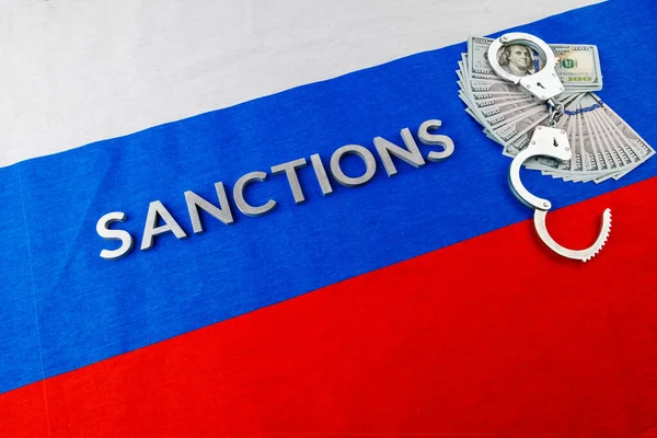 the word sanctions laid with silver metal letters on russian tricolor flag near dollar banknotes and handcuffs in diagonal perspective
