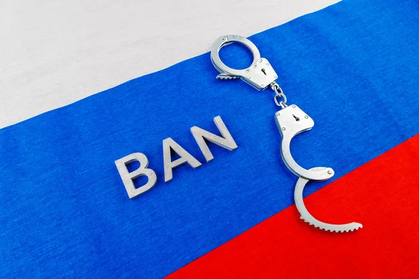 The word ban laid with silver metal letters and handcuffs over fabric Russian Federation flag — Stock Fotó