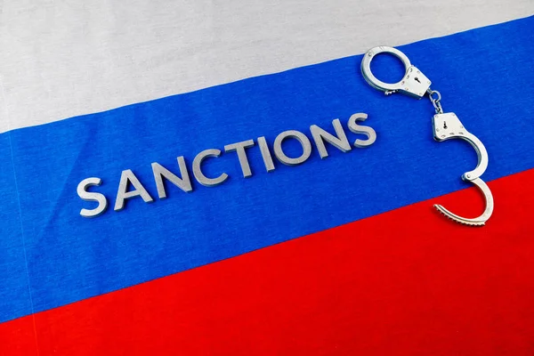 The word sanctions laid with silver metal letters on russian tricolor flag near handcuffs in directly above view — Stok fotoğraf