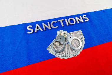 the word sanctions laid with silver metal letters on russian tricolor flag near dollar banknotes and handcuffs in diagonal perspective