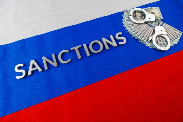 Слова "санкції", закладені срібними металевими літерами на російському триколорному прапорі біля доларових банкнот і наручників в діагональній перспективі — стокове фото