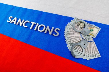 the word sanctions laid with silver metal letters on russian tricolor flag near dollar banknotes and handcuffs in diagonal perspective