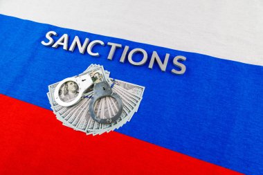 the word sanctions laid with silver metal letters on russian tricolor flag near dollar banknotes and handcuffs in diagonal perspective