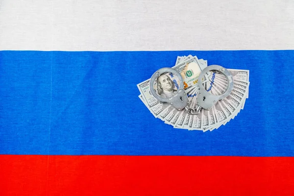 silver metal police handcuffs over paper dollar banknotes of United States of America over national flag of Russian Federation in high angle view