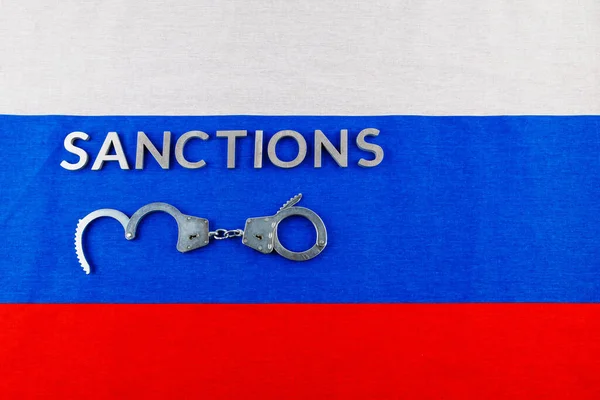 The word sanctions laid with silver metal letters on russian tricolor flag near handcuffs in directly above view — Fotografia de Stock