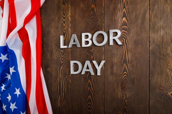 Tahta yüzeyde gümüş metal harflerle, sol tarafında buruşuk Amerikan bayrağıyla...