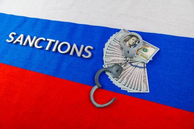 the word sanctions laid with silver metal letters on russian tricolor flag near dollar banknotes and handcuffs in diagonal perspective