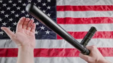 caucasian hands holding black rubber police tonfa baton over blurry US flag