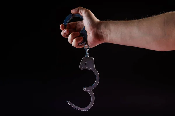 Bare caucasian hand holding opened silver steel handcuffs on black background — Fotografia de Stock