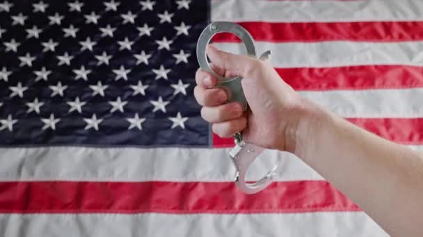 Caucasian hand holding silver metal handcuffs over blurry US flag — Stockvideo