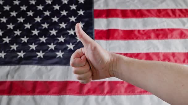 Thumbs up and thumbs down gestures made with caucasian hand in front of blurry US flag — Wideo stockowe