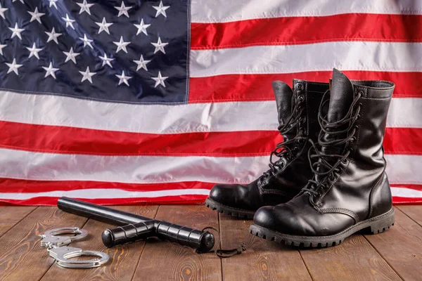 Silver metal handcuffs, black ankle boots and police nightstick near US flag on wooden surface — Foto Stock