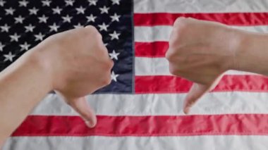 thumbs up and thumbs down gestures made with two caucasian hands in front of blurry US flag