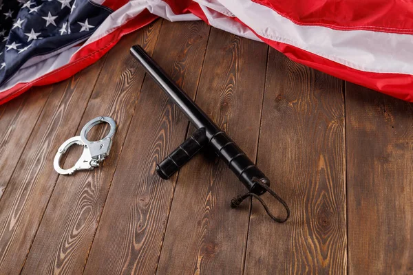 Серебряные металлические наручники и полицейская дубинка возле флага США на деревянной поверхности — стоковое фото