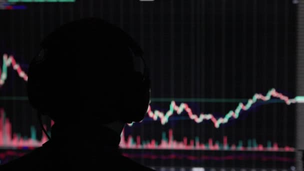 株式市場の取引チャートの前でヘッドフォンをしている男の頭の認識できない黒い背景の上にぼやけた日本の燭台 — ストック動画
