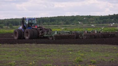 Blue New Holland T9030 tractor with double wheels pulling disc harrow with roller baskets at spring day surrounded with black-headed gulls with slow motion in Tula, Russia - June 4, 2022