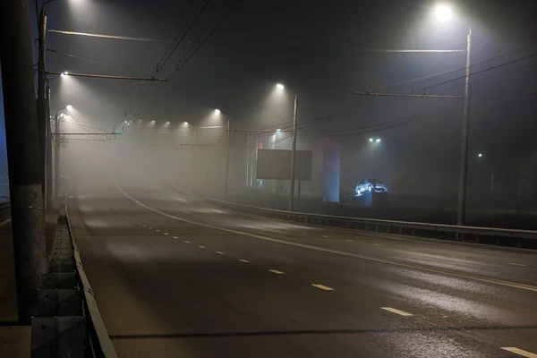 illuminated empty foggy night road with rigid guardrails and car on pedestal near car dealership