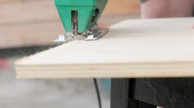 Sawdust flies off a metal blade as the jigsaw cuts into a sheet plywood workpiece. Carpenter cuts off the edge of a plywood plank. Jigsaw cuts into a piece of plywood. Frontal view.