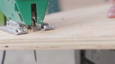 Sawdust flies off a metal blade as the jigsaw cuts into a sheet plywood workpiece. Carpenter cuts off the edge of a plywood plank. Jigsaw cuts into a piece of plywood. Frontal view.