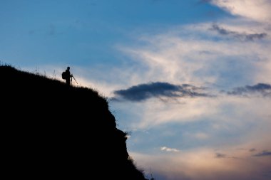 silhouette of unrecognizable photographer with tripod on cliff with evening blue sky with darkening clouds background
