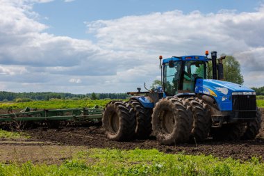 Blue New Holland tractor with double wheels pulling disc harrow with roller basket at hot sunny day in Tula, Russia - June 4, 2022