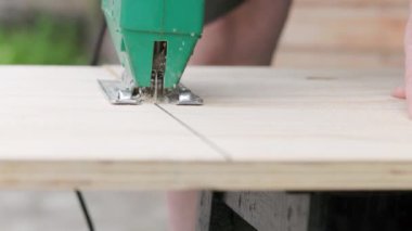 Sawdust flies off a metal blade as the jigsaw cuts into a sheet plywood workpiece. Carpenter cuts off the edge of a plywood plank. Jigsaw cuts into a piece of plywood. Frontal view.