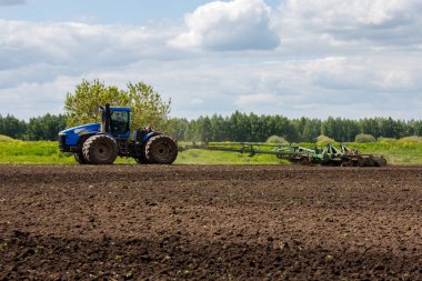 Blue New Holland tractor with double wheels pulling disc harrow with roller basket at hot sunny day in Tula, Russia - June 4, 2022