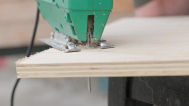 Sawdust flies off a metal blade as the jigsaw cuts into a sheet plywood workpiece. Carpenter cuts off the edge of a plywood plank. Jigsaw cuts into a piece of plywood. Frontal view.