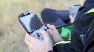 drone operator hands using remote control with sticks and cellphone as monitor at daylight on blurry wild grass background
