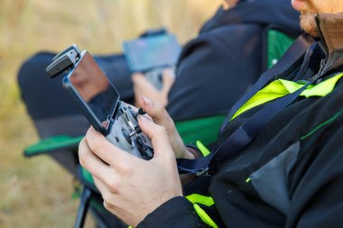 drone operator hands using remote control with sticks and cellphone as monitor at daylight on blurry wild grass background