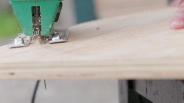 Sawdust flies off a metal blade as the jigsaw cuts into a sheet plywood workpiece. Carpenter cuts off the edge of a plywood plank. Jigsaw cuts into a piece of plywood. Frontal view.