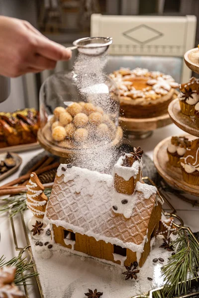女人的手把糖霜撒在姜饼屋上面 有糖果的节庆圣诞餐桌 — 图库照片