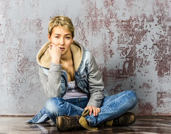 Young beautiful girl with short hair in jeans and a denim jacket sitting at the wooden wall — Stock Photo, Image