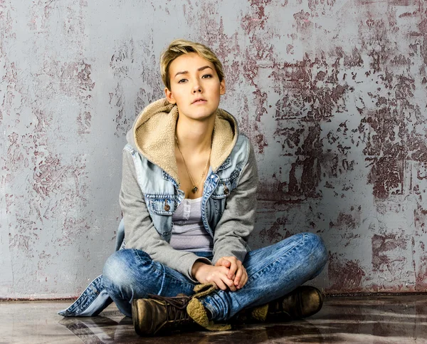 Joven hermosa chica con el pelo corto en jeans y una chaqueta de mezclilla sentado en la pared de madera — Foto de Stock
