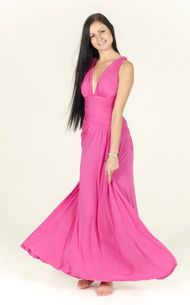 Menina morena bonita com cabelos longos em um vestido rosa com um decote mergulhando pensativamente fica descalça, meio virada Imagem De Stock