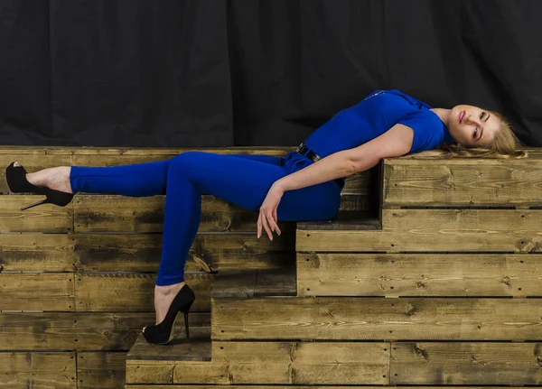 Beautiful long haired blonde in blue overalls and high heels sitting on wooden stairs — Stock Photo, Image