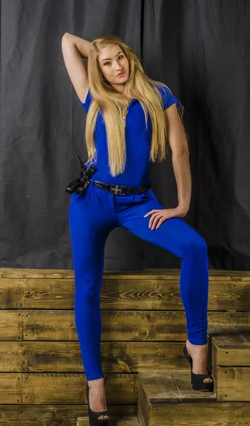 Linda loira de cabelos longos em macacões azuis e saltos altos sentados em escadas de madeira — Fotografia de Stock