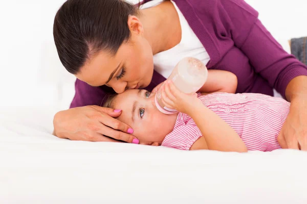 Caring mother kissing baby daughter while she drinking milk — Stock Photo, Image